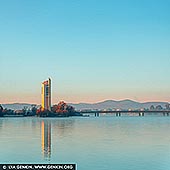 australia stock photography | National Carillon at Sunrise, Canberra, Australian Capital Territory (ACT), Australia, Image ID AU-ACT-CANBERRA-0017. At sunrise, the National Carillon on Queen Elizabeth II Island (formerly known as Aspen Island) emerges in a cinematic palette of teal and orange. The tower's angular Brutalist lines reflect the dawn's glow, standing tall at 50 meters above Lake Burley Griffin. Gifted by Britain in 1970 to commemorate Canberra's 50th anniversary, its 57 bronze bells, cast by John Taylor & Co., span nearly five octaves. The 2019 addition of the Ngunnawal Bell honors the region's Indigenous heritage. As the sun rises, the Carillon's silhouette and resonant chimes offer a serene start to the day.