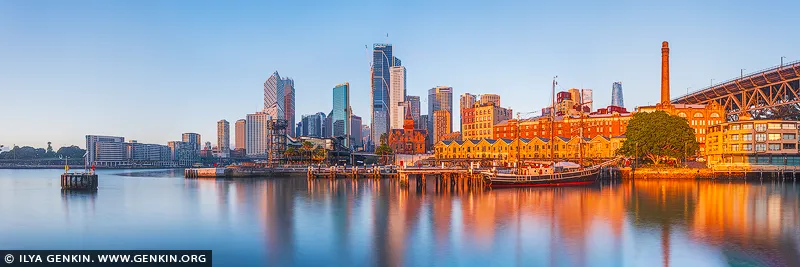 australia stock photography | Golden Stillness, Circular Quay and Sydney City, Sydney, NSW, Australia.. Morning arrives with a rare sense of composure, laying warm amber light across the waterfront while the city rises quietly beyond its glassy reflection. Soft blue water and a clear sky temper the glowing architecture, creating an atmosphere of calm assurance - a presence that brings spaciousness, clarity and an almost meditative stillness into the room. As a design anchor, the artwork commands attention without overwhelming a refined residential or executive interior. Its luminous golds, terracotta warmth and cool harbour blues respond beautifully to changing ambient light, allowing the wall to shift subtly through the day. Presented in a commanding large-format museum presentation, the panoramic proportions truly come alive, extending the visual boundaries of the room and giving the surrounding space a greater sense of breadth and architectural presence.