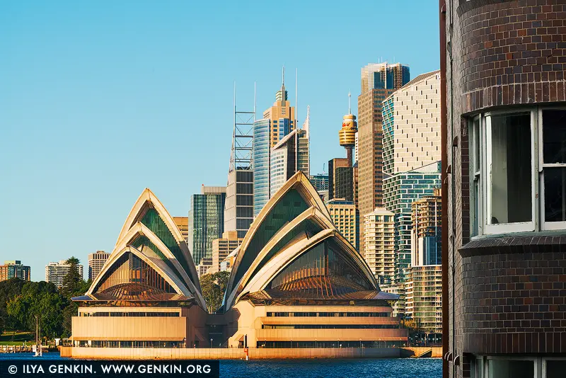 australia stock photography | Golden Light on Icons: Sydney Opera House & Skyline, Sydney, New South Wales (NSW), Australia. Bathed in warm late morning sunlight, the Sydney Opera House stands proudly before the rising geometry of the city's skyline - a captivating dance between heritage and modernity. The glinting sails of the Opera House echo the rhythms of nature, while the angular skyscrapers behind it, including Aurora Place and Chifley Tower, reflect the bold ambition of a contemporary metropolis. Framed by a brick building in the foreground, this intimate cityscape feels both grand and personal - a portrait of Sydney's evolving soul. Ideal for stylish interiors, this artwork adds warmth, culture, and architectural intrigue to any living or professional space.