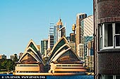 Golden Light on Icons: Sydney Opera House & Skyline - Fine art print of Sydney, New South Wales (NSW), Australia - Bathed in warm late morning sunlight, the Sydney Opera House stands proudly before the rising geometry of the city's skyline - a captivating dance between heritage and modernity. The glinting sails of the Opera House echo the rhythms of nature, while the angular skyscrapers behind it, including Aurora Place and Chifley Tower, reflect the bold ambition of a contemporary metropolis. Framed by a brick building in the foreground, this intimate cityscape feels both grand and personal - a portrait of Sydney's evolving soul. Ideal for stylish interiors, this artwork adds warmth, culture, and architectural intrigue to any living or professional space. - Image ID: AU-SYDNEY-OPERA-HOUSE-0051