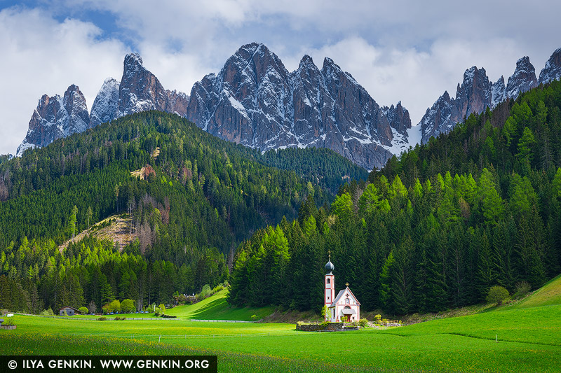 italy stock photography | Church of Saint John, Ranui, Dolomites, Bolzano - South Tyrol, Italy. The Church of Saint John in Ranui stands quietly against the dramatic backdrop of the Dolomites, captured in the golden stillness of a late spring afternoon. The surrounding fields are a lush, vibrant green - alive and full after the thaw - while the jagged mountain peaks above still wear their coat of snow, clinging to winter's final breath. Overhead, dark storm clouds begin to gather, casting a moody contrast between light and shadow, peace and power. The tiny Baroque chapel, with its delicate onion dome and solitary charm, appears almost otherworldly - a steadfast witness to the meeting of seasons and the coming storm.