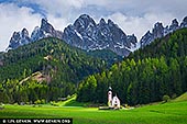  stock photography | Church of Saint John, Ranui, Dolomites, Bolzano - South Tyrol, Italy, Image ID ITALY-DOLOMITES-0001. The Church of Saint John in Ranui stands quietly against the dramatic backdrop of the Dolomites, captured in the golden stillness of a late spring afternoon. The surrounding fields are a lush, vibrant green - alive and full after the thaw - while the jagged mountain peaks above still wear their coat of snow, clinging to winter's final breath. Overhead, dark storm clouds begin to gather, casting a moody contrast between light and shadow, peace and power. The tiny Baroque chapel, with its delicate onion dome and solitary charm, appears almost otherworldly - a steadfast witness to the meeting of seasons and the coming storm.