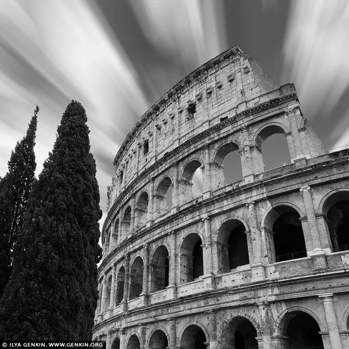 italy stock photography | Timeless Majesty: The Colosseum in Motion, Rome, Lazio, Italy. Swept by streaking clouds and bathed in monochrome drama, this long exposure captures the Colosseum not as a ruin, but as a living monument - defiant, eternal, and etched into the soul of Rome. The ancient arches rise with quiet power, their weathered stones speaking of emperors, crowds, and centuries of spectacle. The tall cypress trees anchor the frame, whispering of Italian elegance and continuity. In black and white, every detail becomes elemental: form, contrast, history, and light. This fine art photograph evokes a sense of grandeur and stillness in motion - ideal for refined interiors seeking a timeless statement of culture and strength.