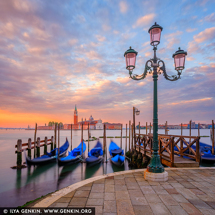 italy stock photography | Venice at Sunrise, Venice, Veneto, Italy. A vibrant sunrise bathes Venice in a kaleidoscope of colours, casting golden, pink, and lavender hues across the sky and shimmering waters. In the background, the elegant silhouette of San Giorgio Maggiore Church stands in serene contrast to the fiery morning light. In the foreground, classic Venetian gondolas gently bob on the lagoon, moored to wooden poles, their dark forms adding depth and rhythm to the tranquil scene. The photograph captures the timeless romance of Venice awakening under a breathtaking dawn.