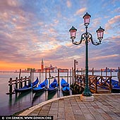  stock photography | Venice at Sunrise, Venice, Veneto, Italy, Image ID ITALY-VENICE-0001. A vibrant sunrise bathes Venice in a kaleidoscope of colours, casting golden, pink, and lavender hues across the sky and shimmering waters. In the background, the elegant silhouette of San Giorgio Maggiore Church stands in serene contrast to the fiery morning light. In the foreground, classic Venetian gondolas gently bob on the lagoon, moored to wooden poles, their dark forms adding depth and rhythm to the tranquil scene. The photograph captures the timeless romance of Venice awakening under a breathtaking dawn.