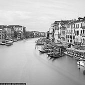  stock photography | Venice Grand Canal, Study 1, Venice, Veneto, Italy, Image ID ITALY-VENICE-0002. Timeless Venice, stripped of color but rich in tone. This black and white long exposure, captured from the iconic Ponte di Rialto, transforms the Grand Canal into a dreamlike flow of light and shadow. The water dissolves into a soft, featureless haze, blurring the passage of time, while the historic facades - from Gothic arches to Renaissance lines - stand in crisp contrast. In daylight's calm, with nearly cloudless skies, the city's elegance emerges in pure form: all texture, rhythm, and geometry. A vision of Venice not as it is, but as it endures.