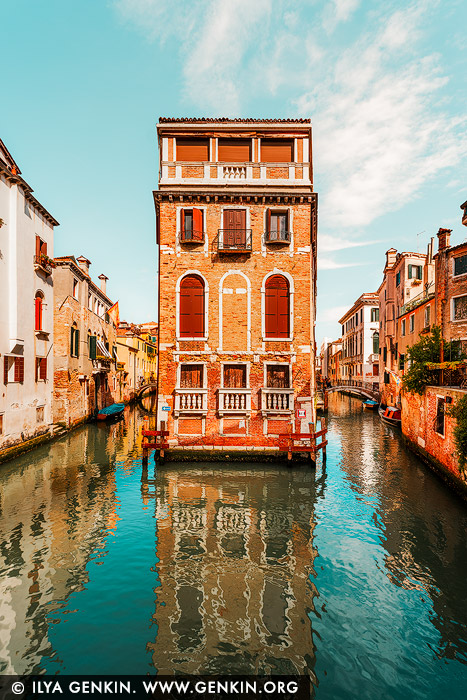italy stock photography | Venice Canals and Streets, Study 2, Venice, Veneto, Italy. Bathed in warm Venetian sunlight, this iconic palazzo stands like a sentinel between two narrow canals, its vibrant terracotta facade glowing against the turquoise waters below. Framed symmetrically by historic buildings, the scene feels almost surreal - like a film set brought to life. The brilliant colours and crisp reflections add a dreamlike intensity, turning a quiet canal into a visual masterpiece. With its bold shutters, elegant arches, and commanding position, the building becomes a symbol of Venice itself: graceful, storied, and impossibly beautiful.