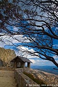 landscapes stock photography | Old Stone Hut and View over Australian Alps from The Horn, Mount Buffalo National Park, Victoria, Australia, Image ID MOUNT-BUFFALO-VIC-0008. Vertical image of the old rock built hut nicely surrounded by a curved dry tree at the starting point of the Horn mountain summit walking in Mount Buffalo National Park, Victoria Hight Country, Australia. This viewpoint offers great view over Australian Alps for all tourists and visitors.