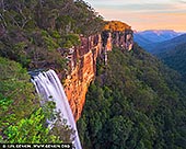 Fitzroy Falls at Sunset - Fine art print of Morton National Park, NSW, Australia - The last light of the day rests on the sandstone cliffs, turning their edges warm against the deep green of the valley below. Fitzroy Falls spills over the escarpment in a steady white rush, its movement set against the quiet strength of rock, forest and distant blue ridgelines. There is a sense of height here, but also stillness - the kind found only when a landscape is caught between daylight and dusk. This image carries the feeling of standing at the edge of something vast. The falls, the cliffs and the layered wilderness beyond evoke power without noise, offering a moment of escape that feels grounded and expansive. It is a scene of contrast: motion and stillness, warmth and shadow, the intimacy of water beside the open distance of Morton National Park. On a wall, this print brings depth, natural presence and a calm sense of scale. Its warm sunset tones and rich greens make it well-suited to spaces where you want the room to feel settled, considered and connected to the landscape - from a living room to a quiet office or studio. Offered as a fine art wall art print, created to be lived with beyond the screen. - Image ID: AU-FITZROY-FALLS-0001