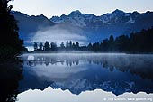landscapes stock photography | Dawn at Lake Matheson, Lake Matheson, South Westland, South Island, New Zealand, Image ID NZ-LAKE-MATHESON-0003. Morning fog rising above Lake Matheson and Mount Cook and Mount Tasman reflect in the lake near Fox Glacier, Westland, Tai Poutini National Park, South Island, New Zealand. Lake Matheson is an ideal location for the scenic picture postcard. It's a small lake surrounded by trees. That means it is almost always has still, flat water surface - exactly what you need for reflections. It also has a view out over the tallest section of the New Zealand's Southern Alps - with Mt Cook (Aoraki) and Mt. Tasman. So it's the perfect spot for a nice picture of the distant snow-capped mountains with reflection in the still water.