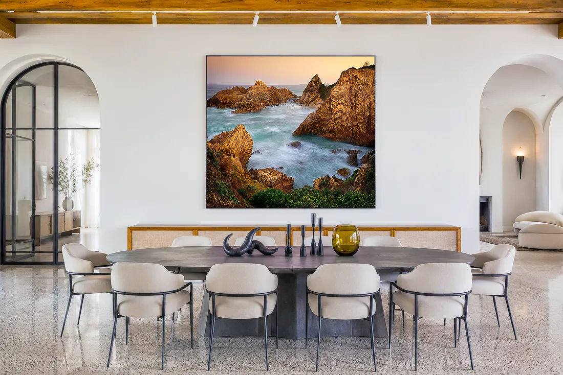 Dining room in a luxury with a large coastal landscape photograph hung above a sideboard behind a long dining table.